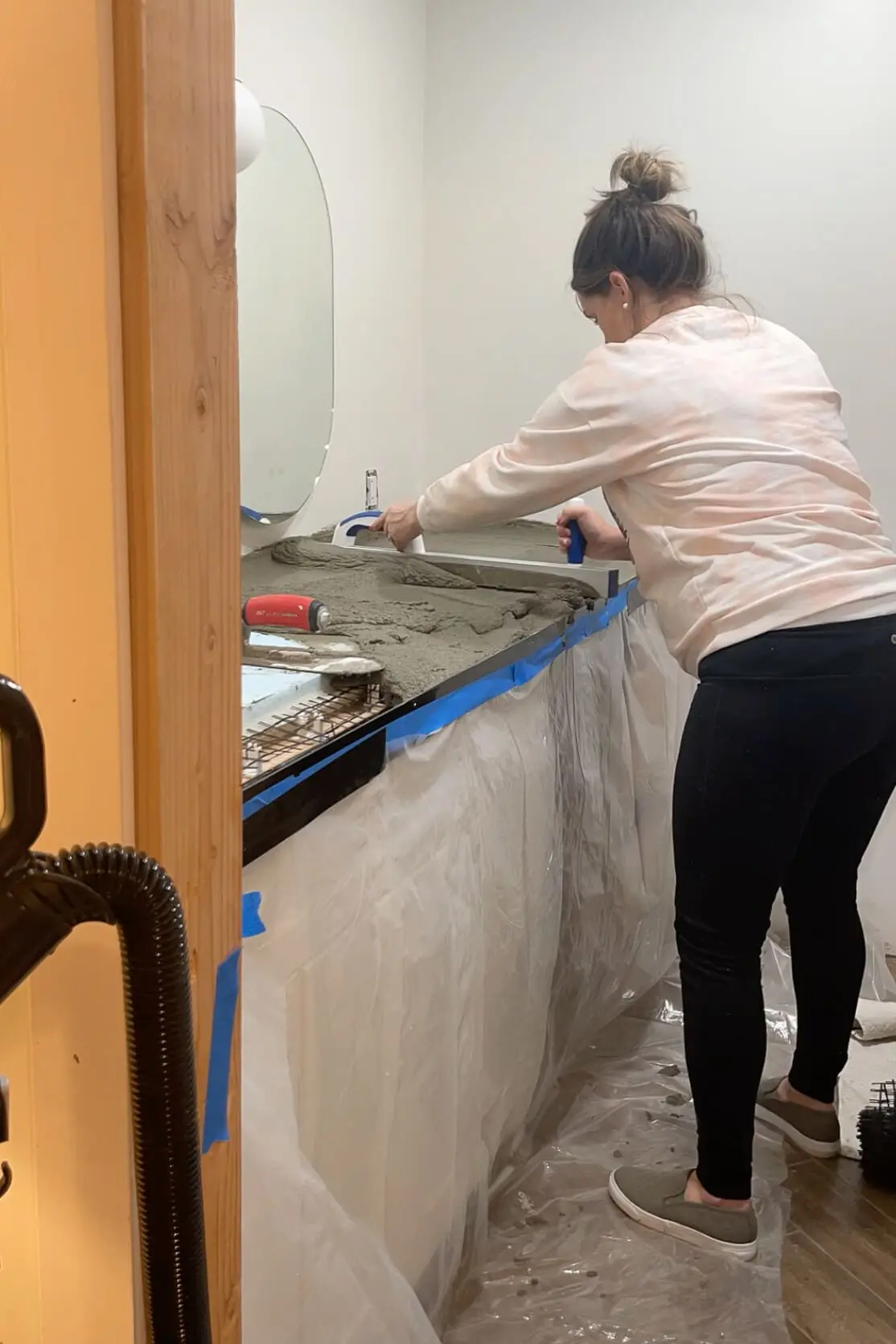 A woman leveling wet concrete on a countertop mold with a screed tool. The workspace features a lined plastic barrier, a black plastic frame, and an oval mirror on the wall, with tools resting on the edge of the mold.